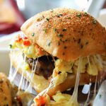 A close-up of breakfast sliders featuring layers of sausage, bacon, egg, and melted cheese sandwiched between golden brown slider buns, arranged neatly on a white platter.